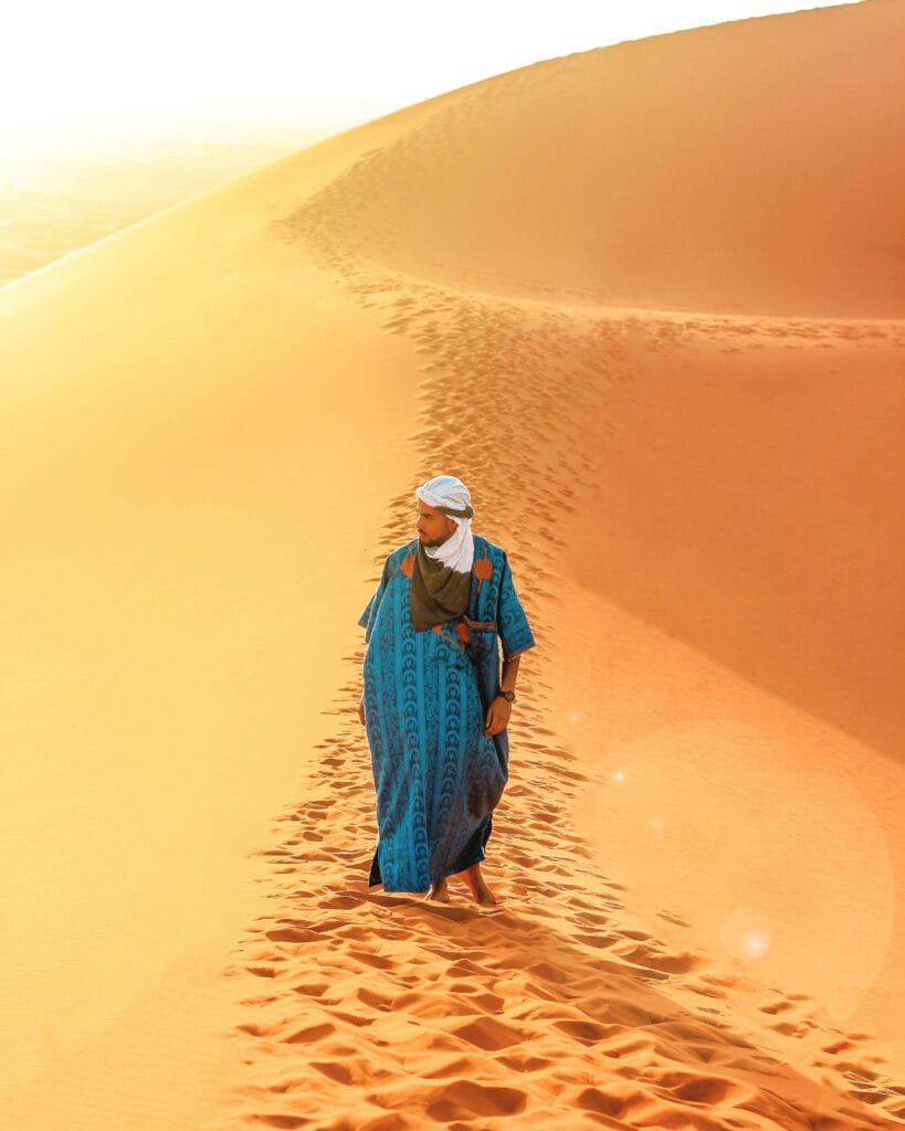 A person wearing traditional clothing walks across the sand dunes of the Sahara Desert at sunrise.